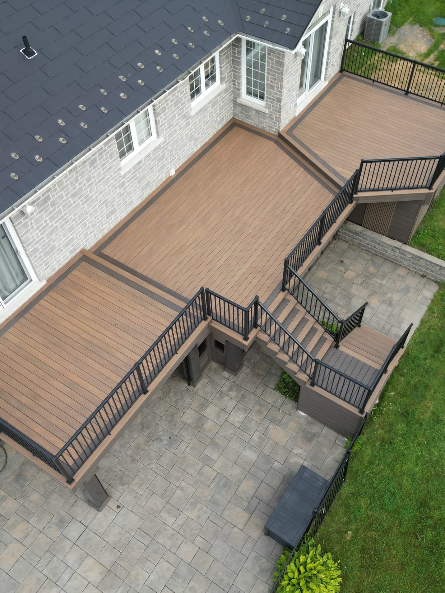 Aerial view of a large multi-level composite deck with black aluminum railing over a paver patio
